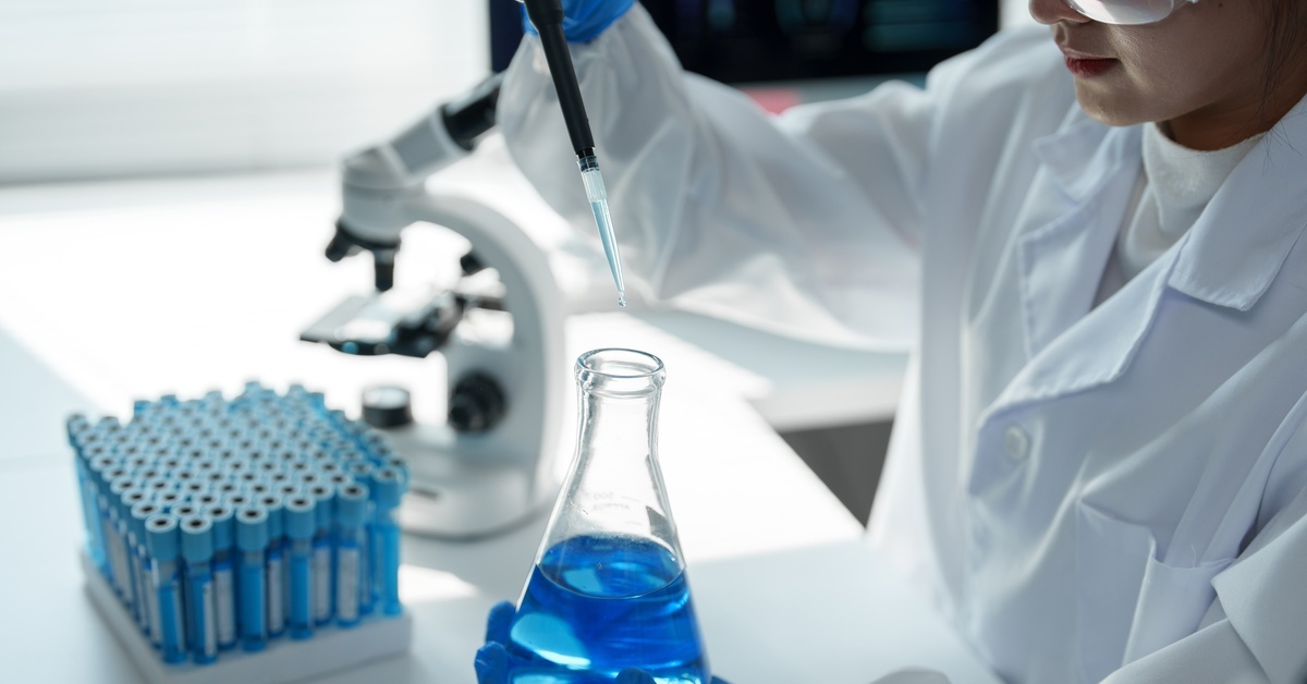 A scientist holding a large pipette and a glass container with blue liquid next to a test tube rack and microscope on a desk.