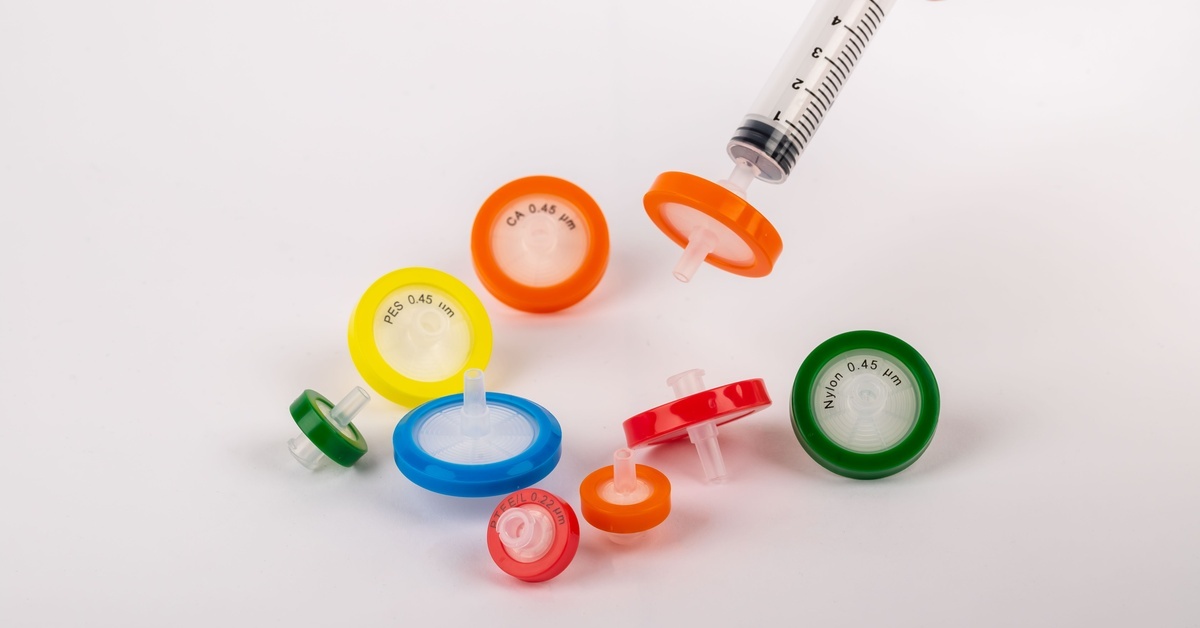 Multiple syringe filters with different color rubbers placed over a white surface with a hand holding a syringe.