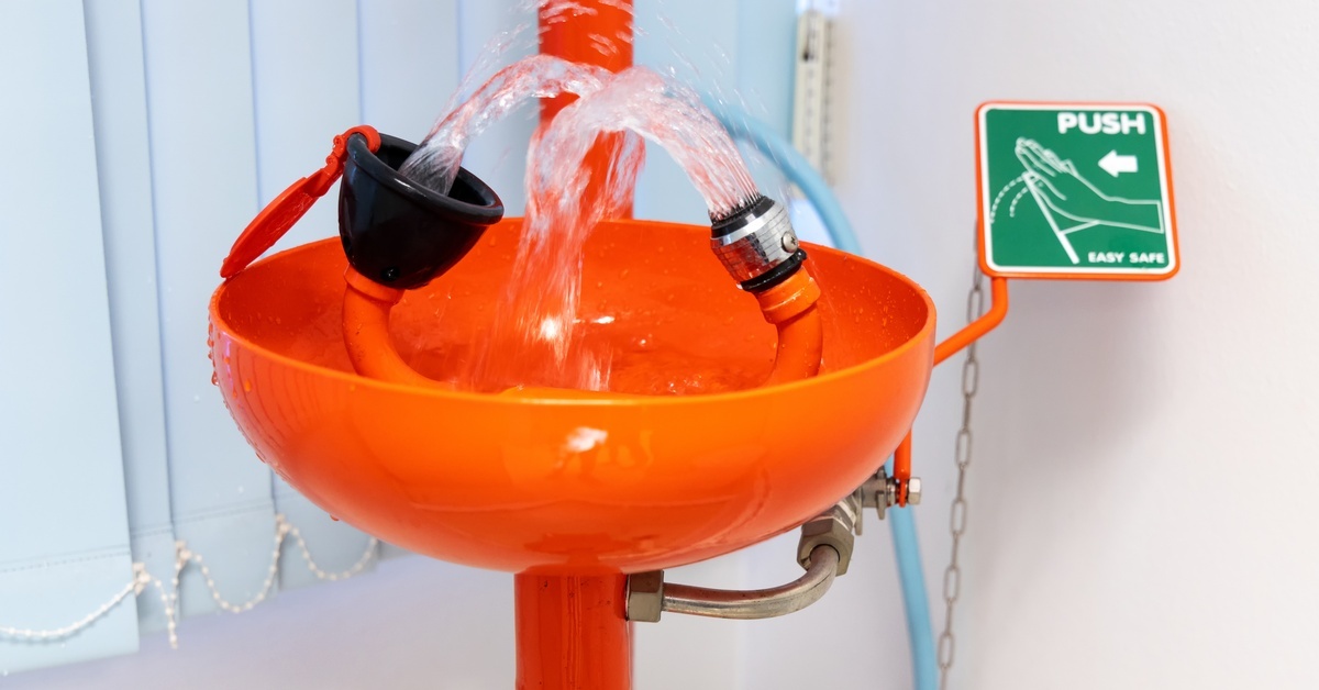 An orange emergency eyewash station with water spouting from both nozzles and a green sign instructing users to push a lever.