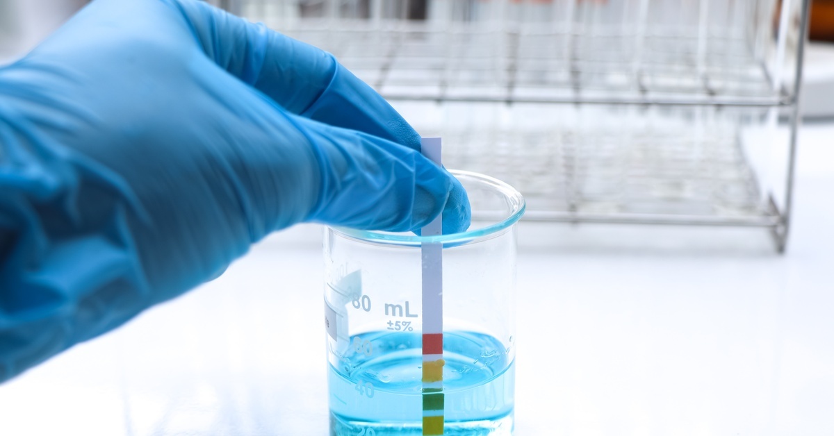 A person wearing a blue latex glove holds a paper stick with multiple colors on its end inside a beaker full of blue fluid.