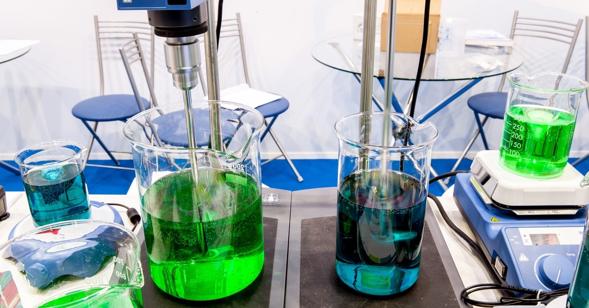 Multiple beakers full of green and blue liquids, with multiple stirring machines and table and chairs surrounding them.