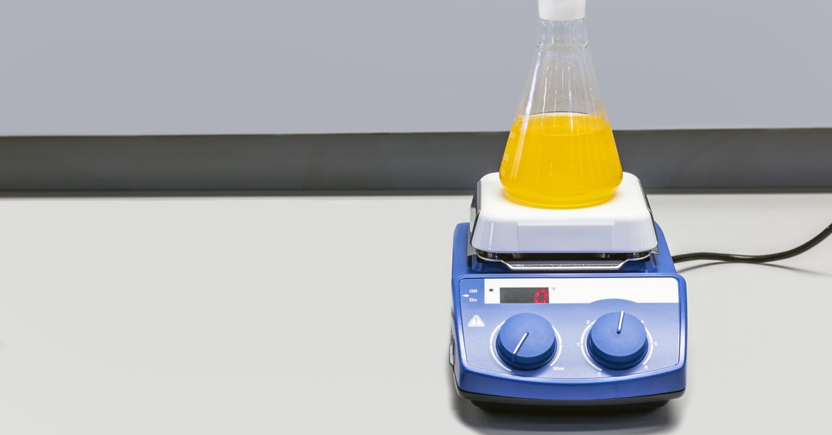A machine with knobs on a white surface, with a chemical flask full of a yellow liquid placed on top.