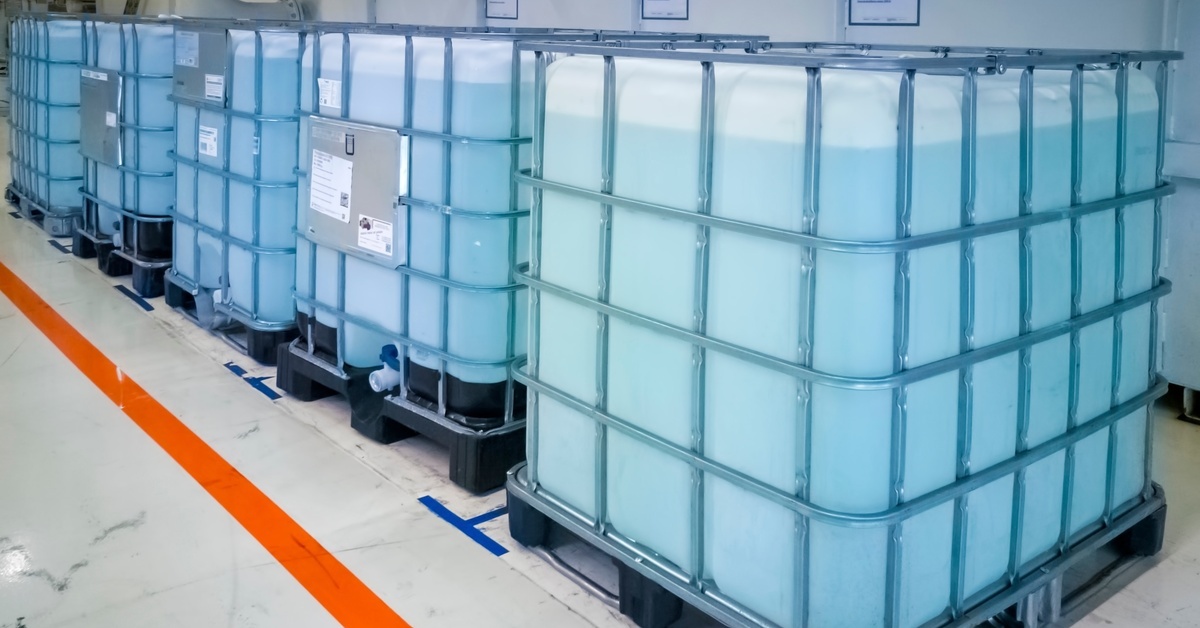 A line of cubic, plastic chemical containers in an industrial space each filled with a light blue liquid.