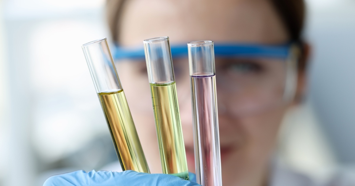 A woman wearing goggles and gloves holds three test tubes filed with liquids that darken in color going from right to left.